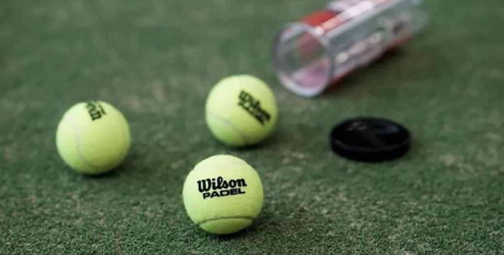 Tre gule padel bolde med sort wilson logo ligger på en grøn måtte med et gennmsigtig padel bold rør ved siden af, hvor det sorte låg er taget af og ligger ved siden af